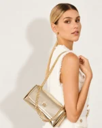 Plano médio da modelo feminina de semiperfil de costas virando o rosto para a câmera com a bolsa tiracolo dourada no ombro.