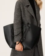 Plano detalhe do torso da modelo com casaco cinza segurando a bolsa tote preta à frente do corpo.