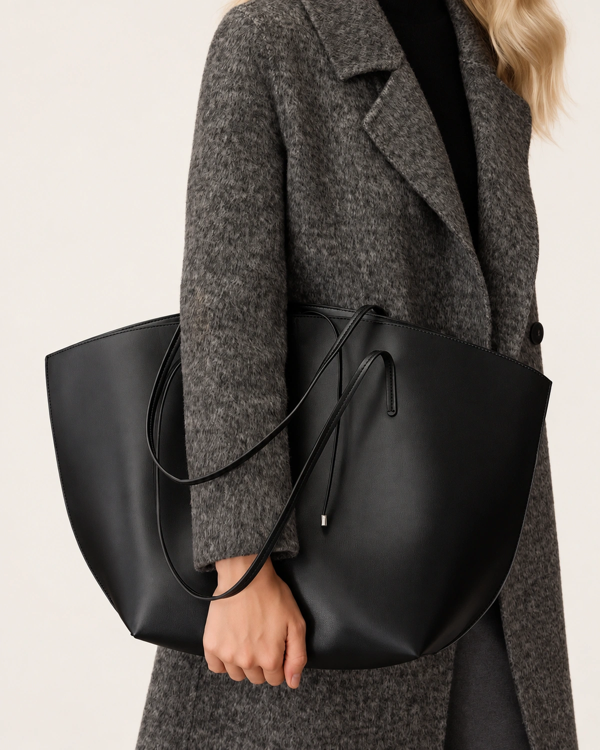 Plano detalhe do torso da modelo com casaco cinza segurando a bolsa tote preta à frente do corpo.