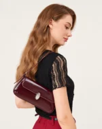 Plano médio da modelo demonstrando a bolsa tiracolo bordeaux suspensa sob o ombro em vista posterior e perfil.