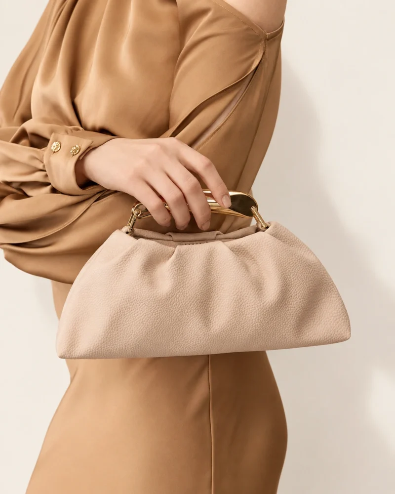 Bolsa Satchel Drapeada com Alça Escultural Metálica em tom areia com alça escultural metálica.