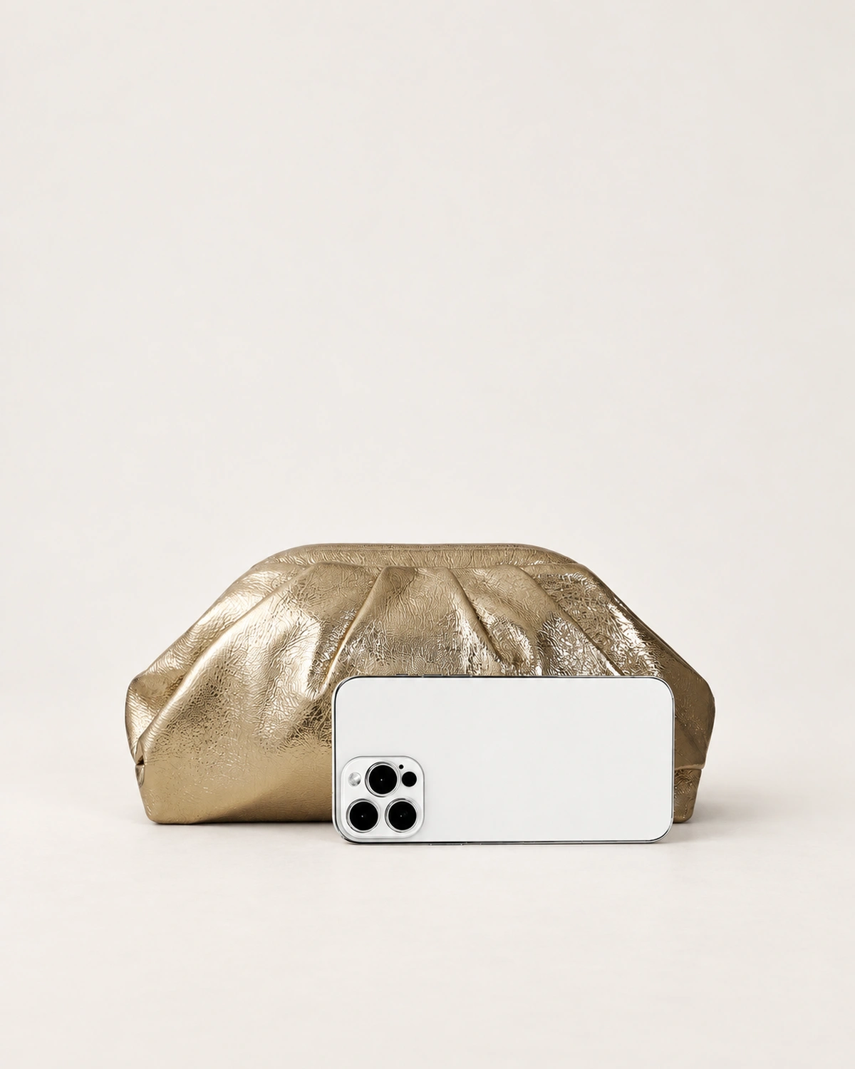 Comparativo de proporção e tamanho real da bolsa clutch drapeada dourada ao lado de smartphone no estúdio mineral.
