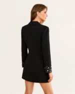 Costas e perfil da modelo com vestido blazer alfaiataria preto acinturado e punhos com pérolas.