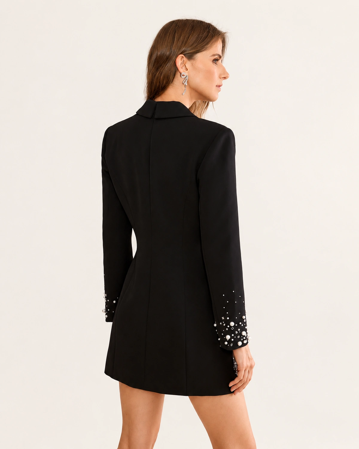 Costas e perfil da modelo com vestido blazer alfaiataria preto acinturado e punhos com pérolas.