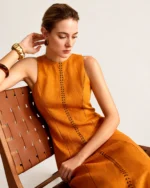 Editorial com modelo sentada em cadeira de design exibindo o corte e fluidez do vestido na cor Terracota Solar.